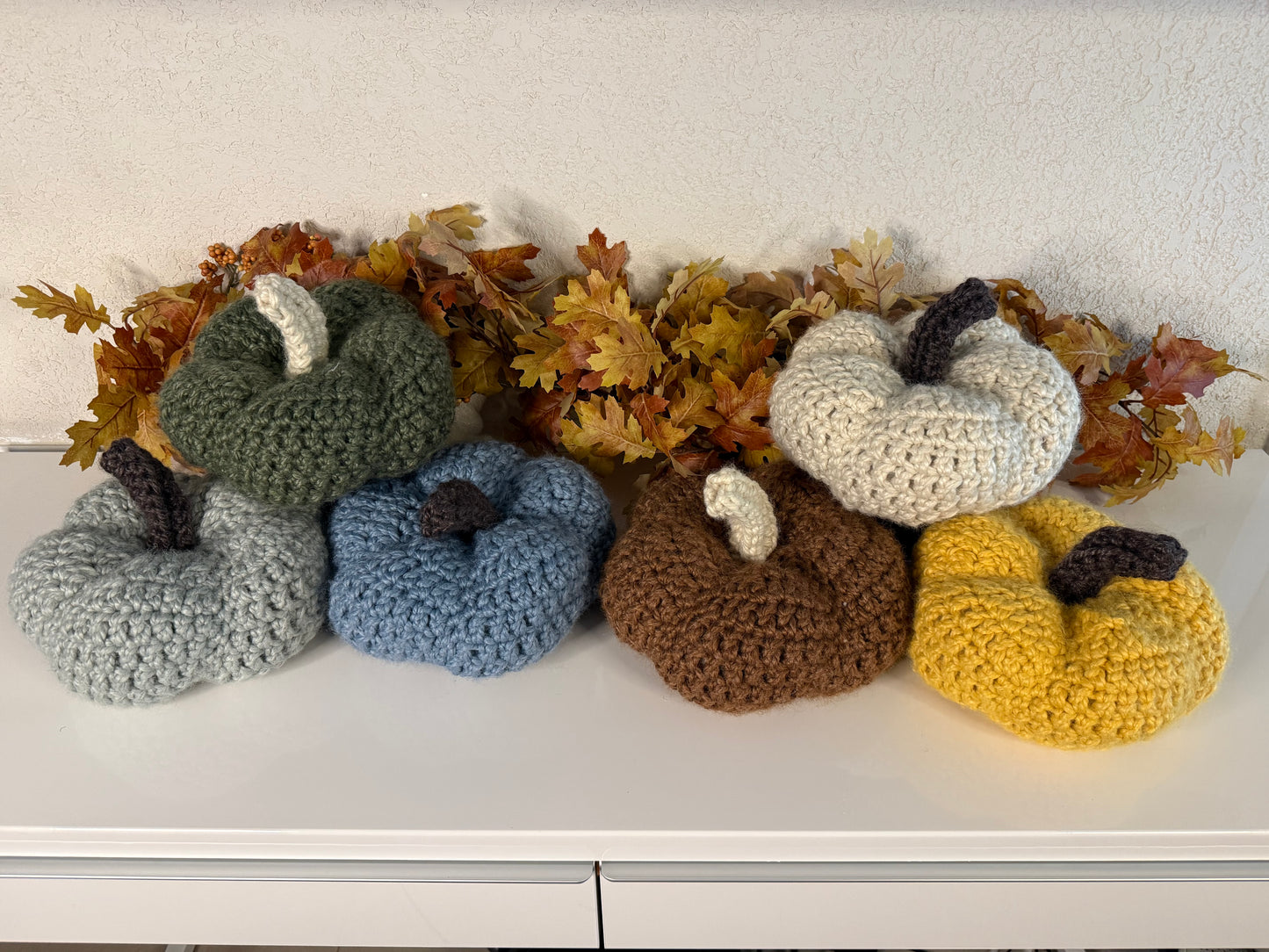 Set of crocheted pumpkins in various colors on a white surface with autumn leaves.