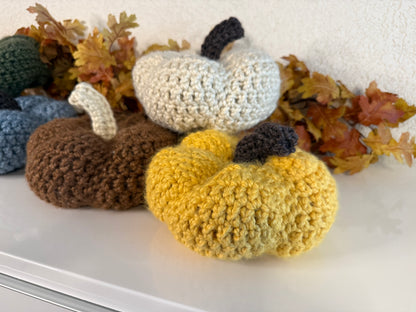 Crocheted pumpkins in various colors on a white surface with autumn leaves.